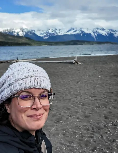 Kathy standing on the shoreline near Glacier Point with snowcapped mountains in the background after arriving from Skagway.
