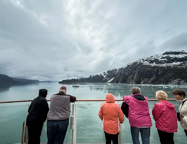 Cruise passengers stand along the deck railing during an Alaska cruise, looking out at calm water, floating ice, and snow-covered mountains.