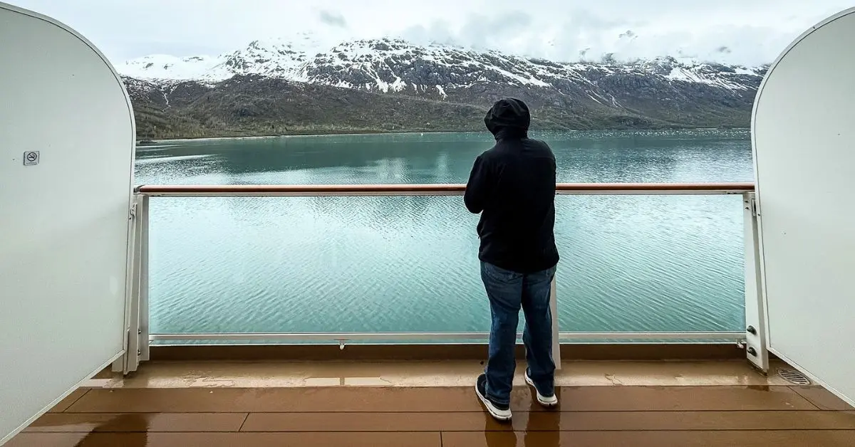 featured image: Person (hubby) standing on a cruise ship balcony overlooking calm water and snow-covered mountains, showing why I’d still splurge on a balcony cabin for an Alaska cruise.