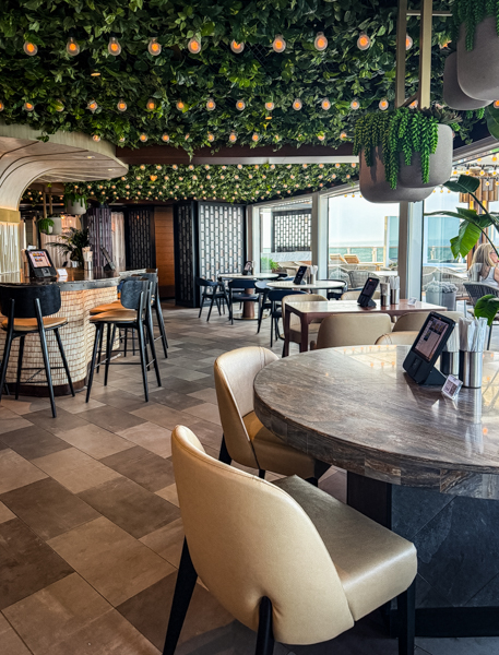 Seating area inside Indulge Food Hall on Norwegian Aqua with ordering tablets, greenery, and ocean views.