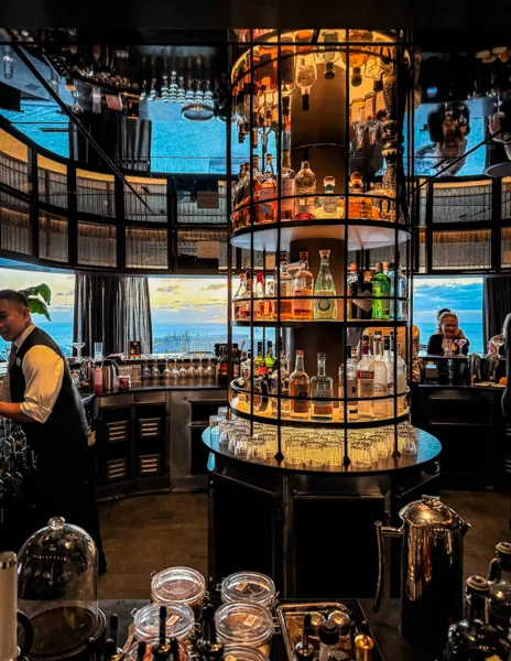 Norwegian Aqua The Haven Bar at sunset with illuminated liquor display, bartenders, and ocean views.