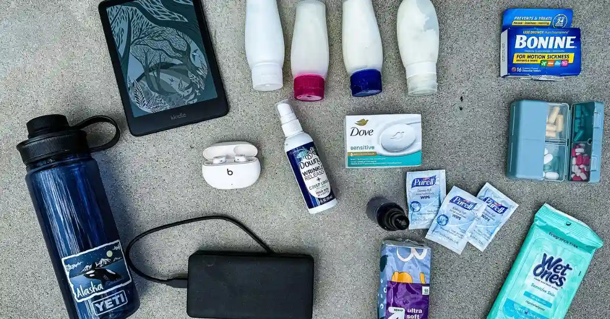 Cruise survival kit essentials laid out with a Kindle, reusable water bottle, portable charger, toiletries, seasickness medicine, medications, wrinkle release spray, sanitizing wipes, and earbuds.