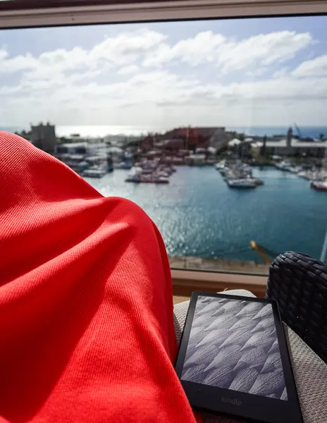 Kindle resting on a cruise ship balcony beside a red dress, with a marina and ocean view in the background.