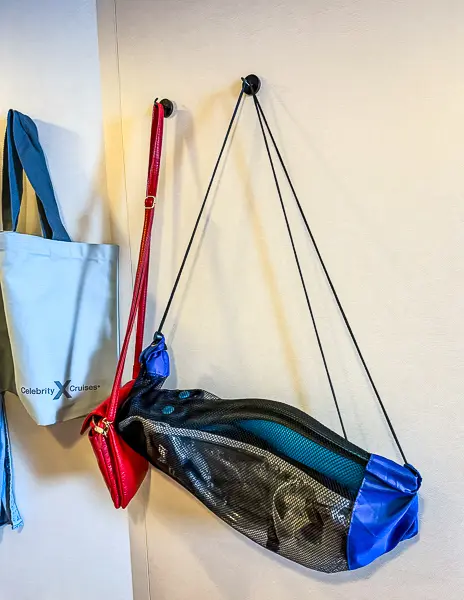 Magnetic hooks on a cruise cabin wall holding a tote bag, red purse, and lightweight travel bag.
