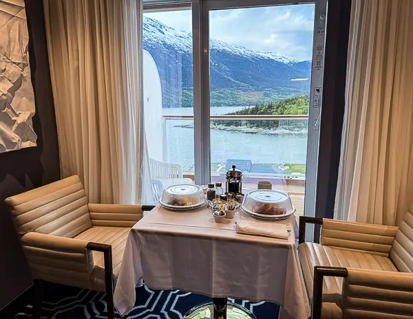 NCL Haven butler breakfast setup with a white tablecloth and covered dishes in the suite, overlooking the mountains from the balcony.