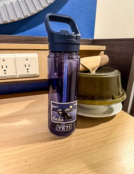 Blue YETI reusable water bottle with an Alaska sticker sitting on a cruise cabin desk beside electrical outlets.