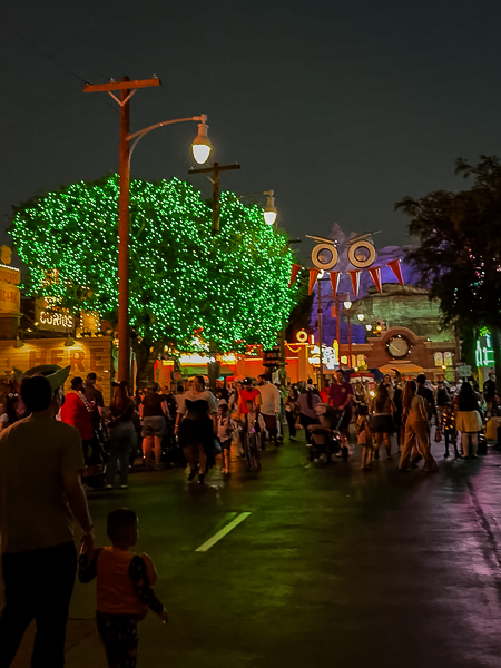Guests walking through Cars Land at Disney California Adventure during Oogie Boogie Bash, with Halloween decorations and trees illuminated in green lights.