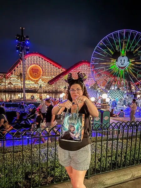 Holding up my Oogie Boogie Bash treat bag at night with Pixar Pier and the illuminated Mickey Ferris wheel in the background.